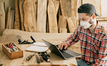 男人戴着面具在笔记本电脑在木桌上木工工具。